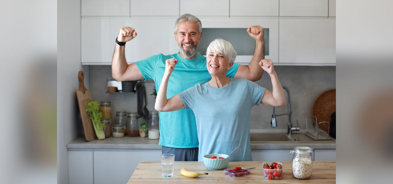 A man and woman in casual workout attire flex their biceps behind a kitchen island that is topped with fruit and oatmeal