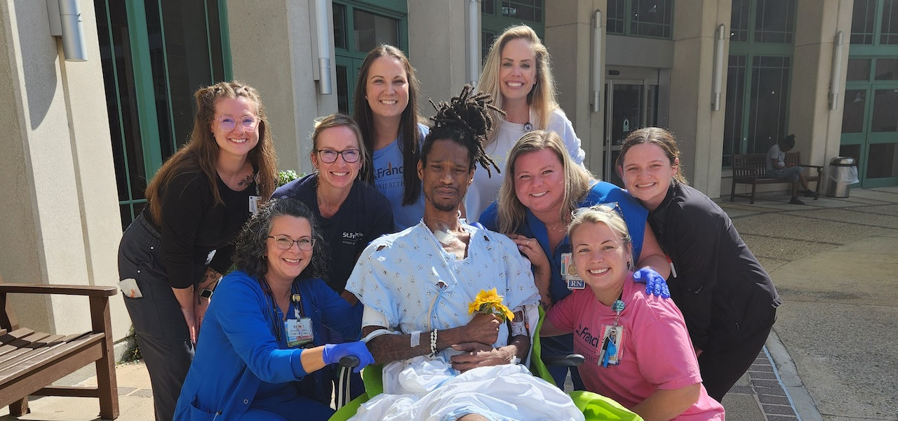 Photograph of a diverse group of nine healthcare professionals and one patient in a hospital outdoor area. The patient, a Black man with short dreadlocks, sits in a wheelchair wearing a light blue hospital gown with white patterns.